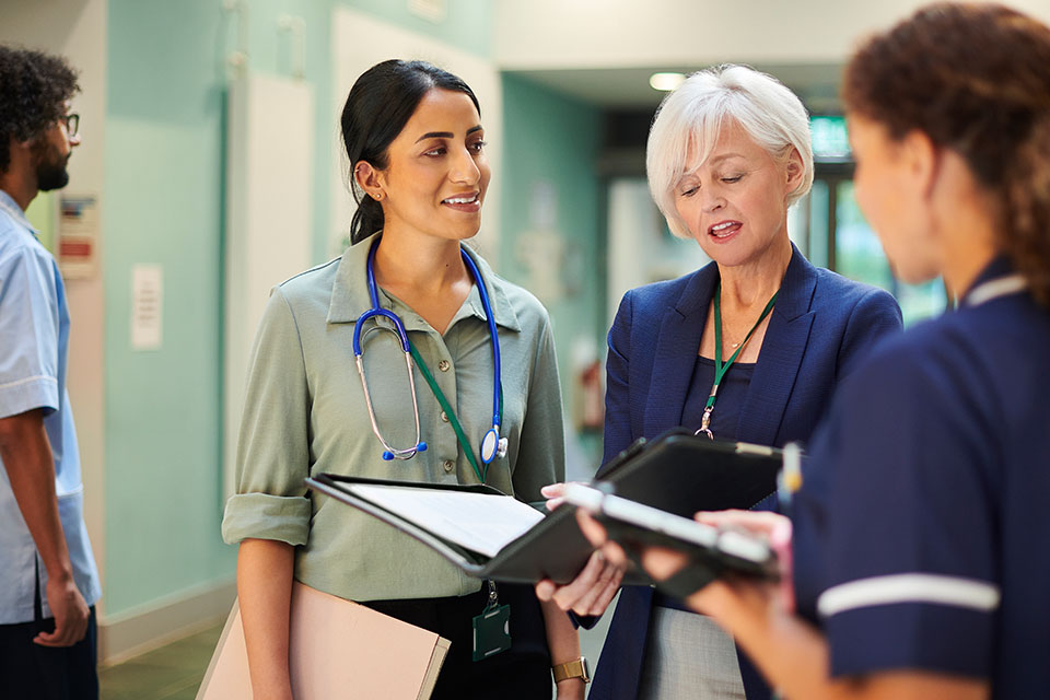 Three healthcare providers (a nurse, doctor, and healthcare administrator) discussing work in a hospital hallway.