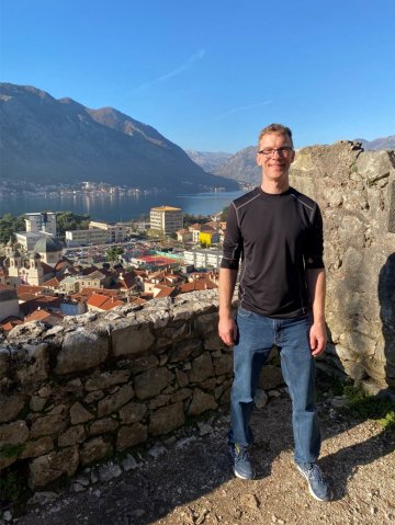 Luke Perry stands in front of a small stone wall in Montenegro with houses and buildings below.