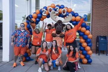 Students in orange t-shirts stand with Trax the Moose under an orange and blue balloon arch.