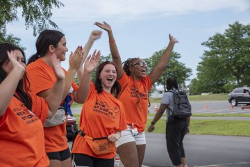 Students in orange t-shirts happily shout and wave their arms to greet newcomers to campus.